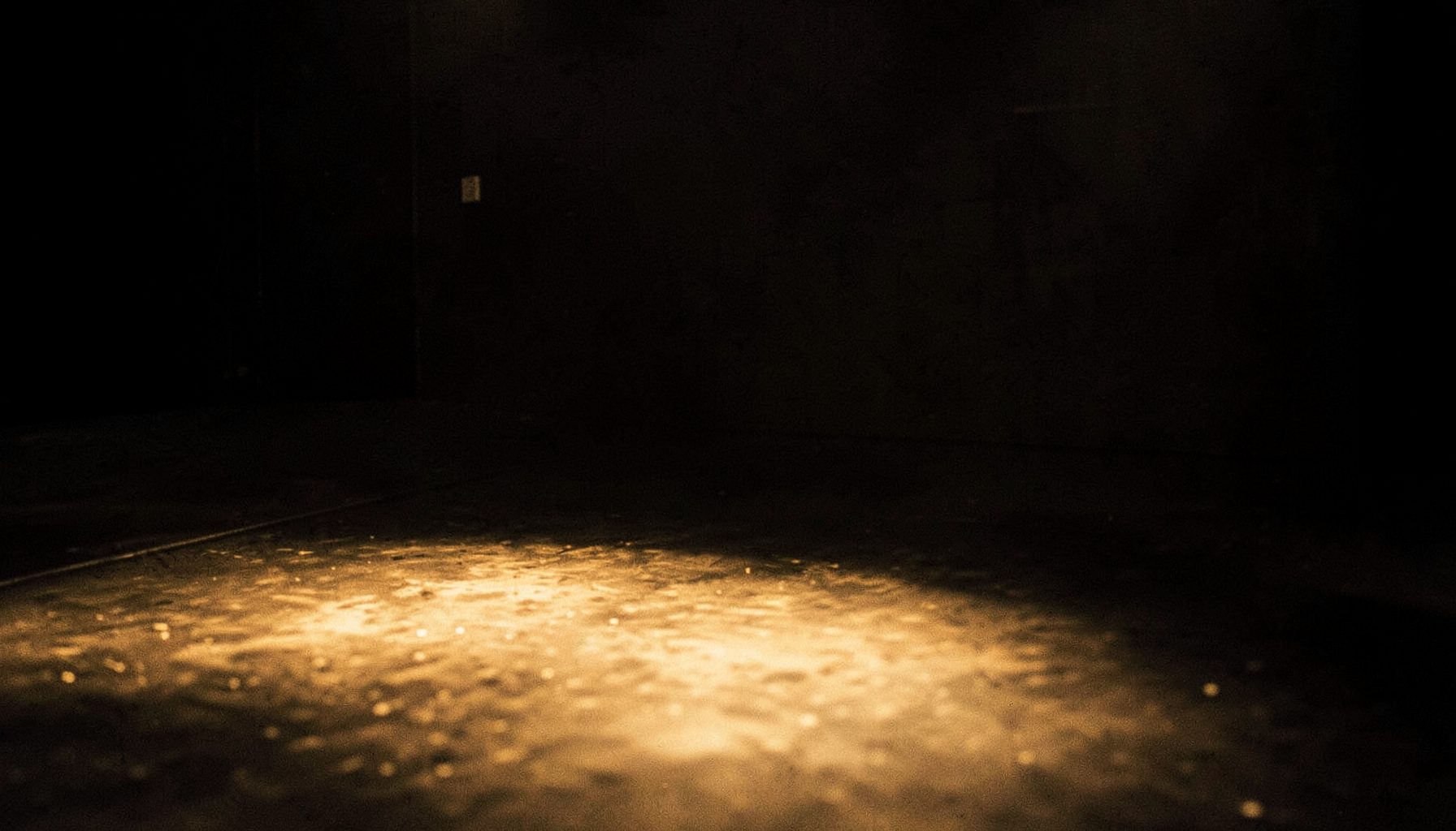 A close, textured view of a performance space with deep shadows and soft golden light pooling on a bare floor, suggesting movement and aftermath.