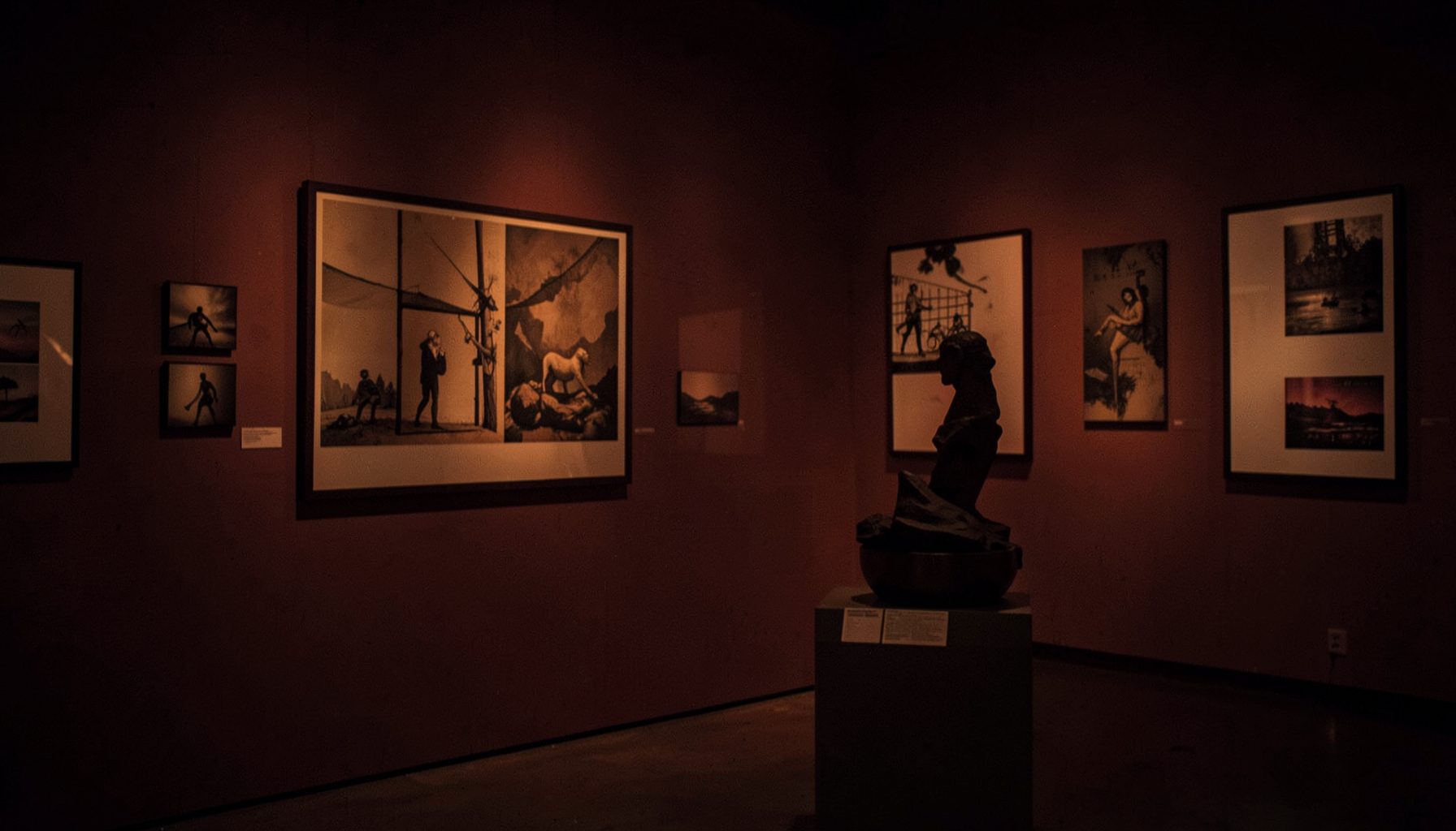 A dimly lit gallery wall with framed artworks and a sculptural piece on a plinth, warm amber and deep burgundy tones, quiet and contemplative atmosphere