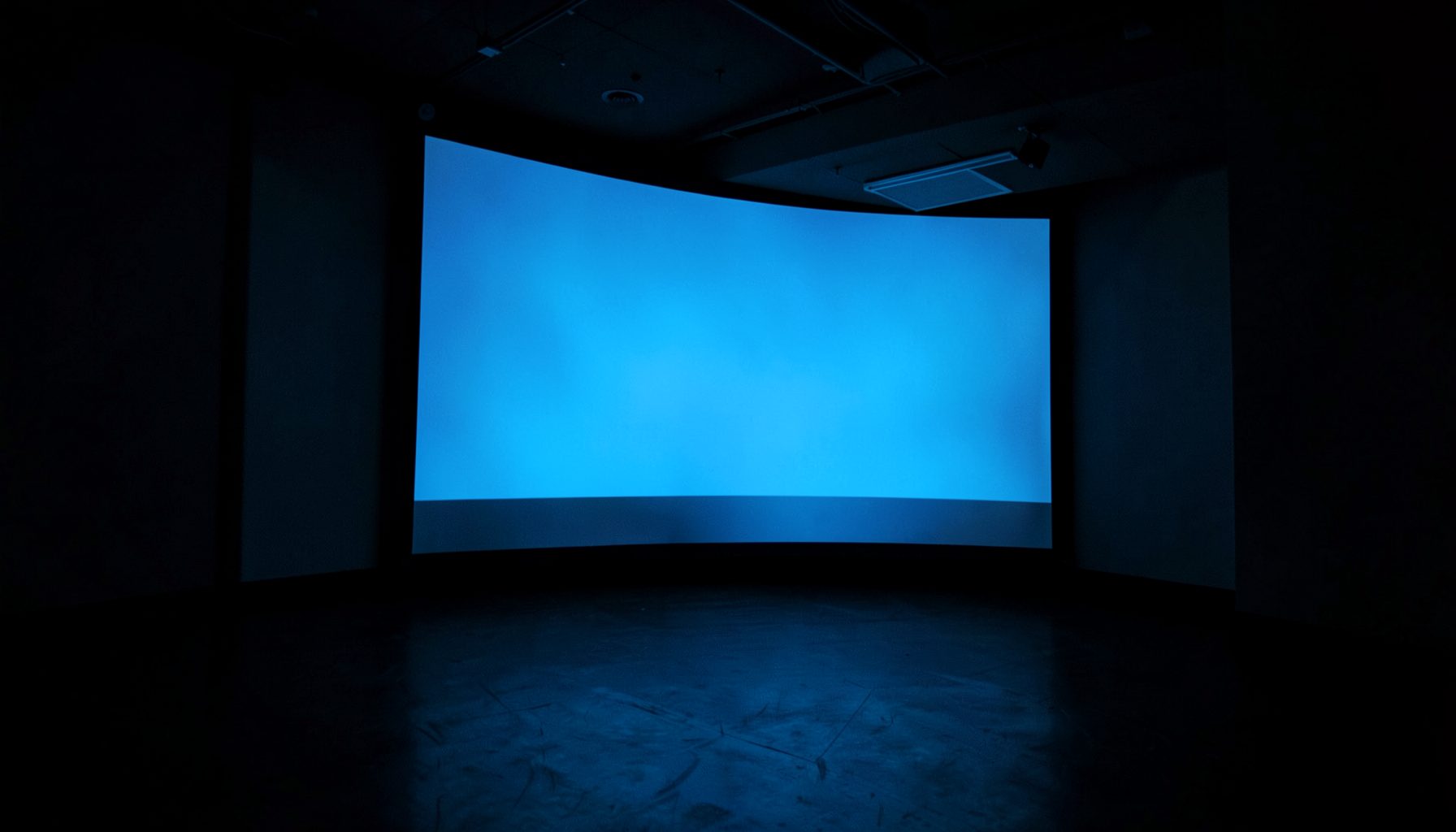 A dimly lit projection room with a large screen glowing in cool blue tones, casting soft reflections across a dark floor; the atmosphere is quiet and contemplative.