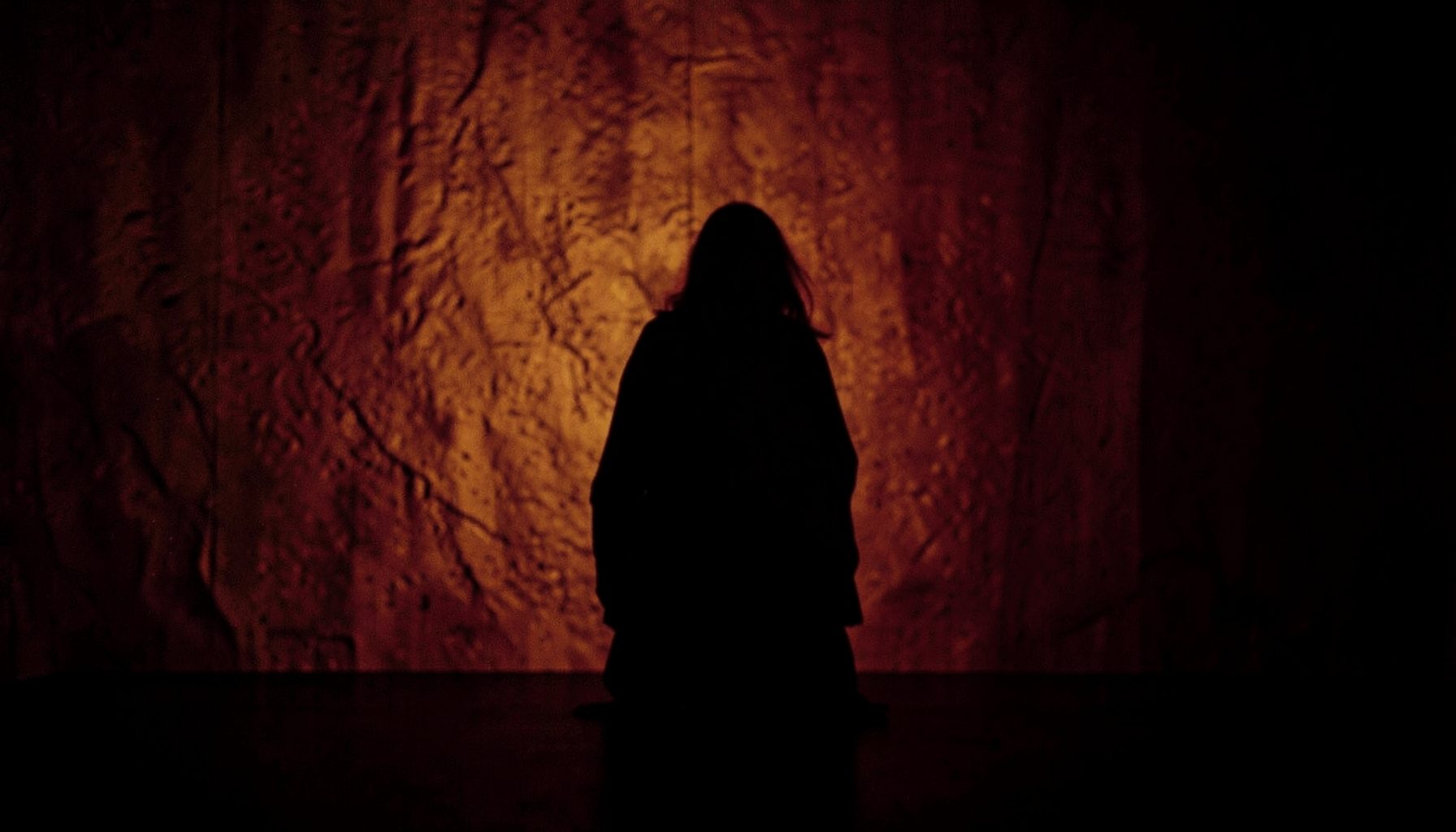 A dimly lit stage with warm amber and deep burgundy tones, a lone figure in silhouette against a textured backdrop, evoking intimacy and raw presence.