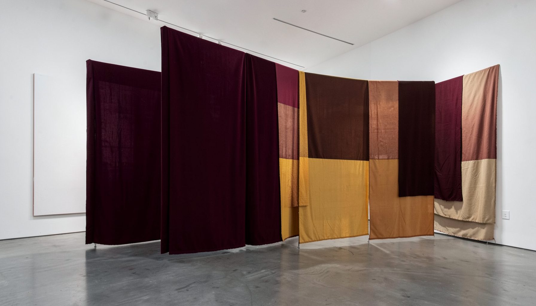 A large-scale gallery installation with layered fabric panels in deep burgundy and ochre, softly lit against white walls