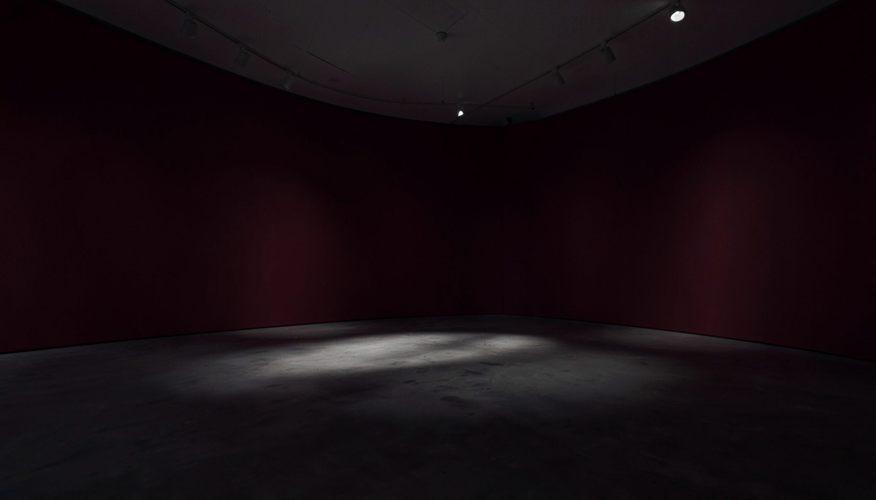 A wide, atmospheric installation view in deep burgundy and charcoal tones, with soft gallery lighting casting long shadows across an open floor, evoking stillness and contemplation.