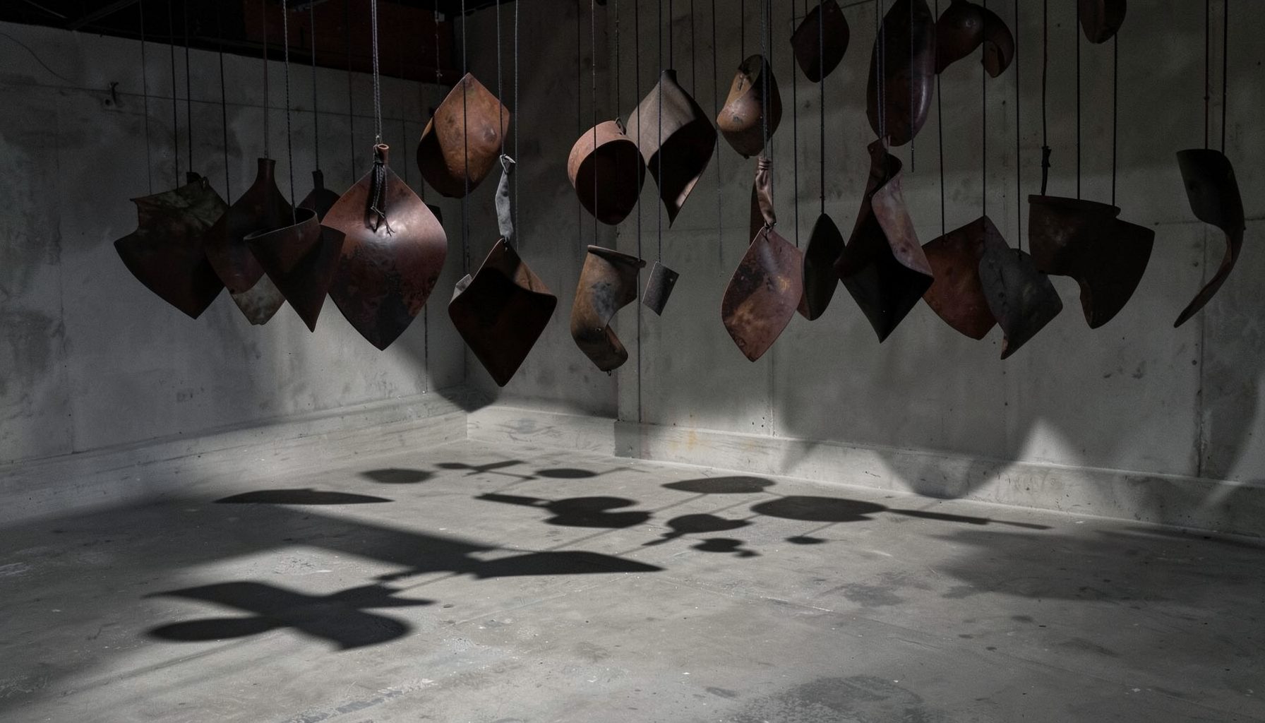 An installation with suspended sculptural forms in dark earth tones, casting long shadows across a concrete floor